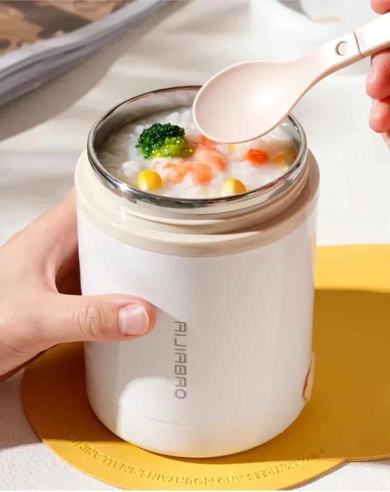 Person holding a white thermos with a lid, containing a bowl of soup with vegetables, on a yellow coaster.