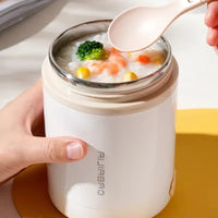 Person holding a white thermos with a lid, containing a bowl of soup with vegetables, on a yellow coaster.