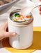 Person holding a white thermos with a lid, containing a bowl of soup with vegetables, on a yellow coaster.